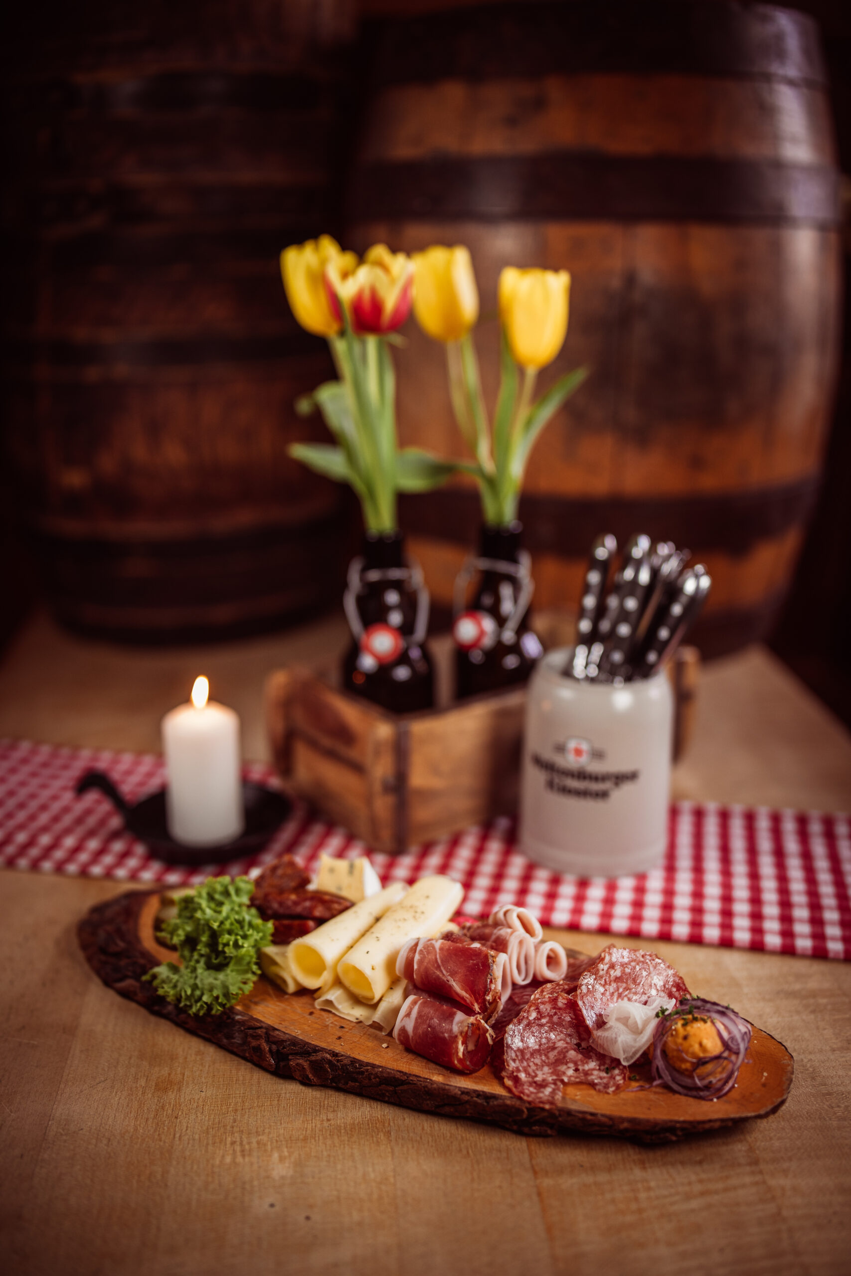 Ein rustikales Holzbrett, reich belegt mit verschiedenen Wurst- und Käsespezialitäten. Man sieht gerollten Käse, geräucherten Schinken, verschiedene Salamisorten, rote Zwiebelringe und ein Salatblatt. Ein rot-weiß kariertes Tuch ist im Hintergrund.