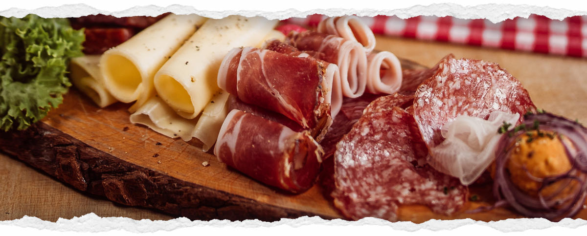 Ein rustikales Holzbrett, reich belegt mit verschiedenen Wurst- und Käsespezialitäten. Man sieht gerollten Käse, geräucherten Schinken, verschiedene Salamisorten, rote Zwiebelringe und ein Salatblatt. Ein rot-weiß kariertes Tuch ist im Hintergrund.