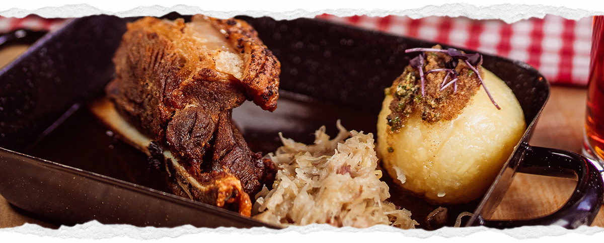 Ein traditioneller Schweinshaxe in einer schwarzen Gusspfanne, daneben Sauerkraut und ein Kartoffelkloß mit Kräutern. Im Hintergrund ist ein rot-weiß kariertes Tuch und ein dunkles Bierglas zu sehen.
